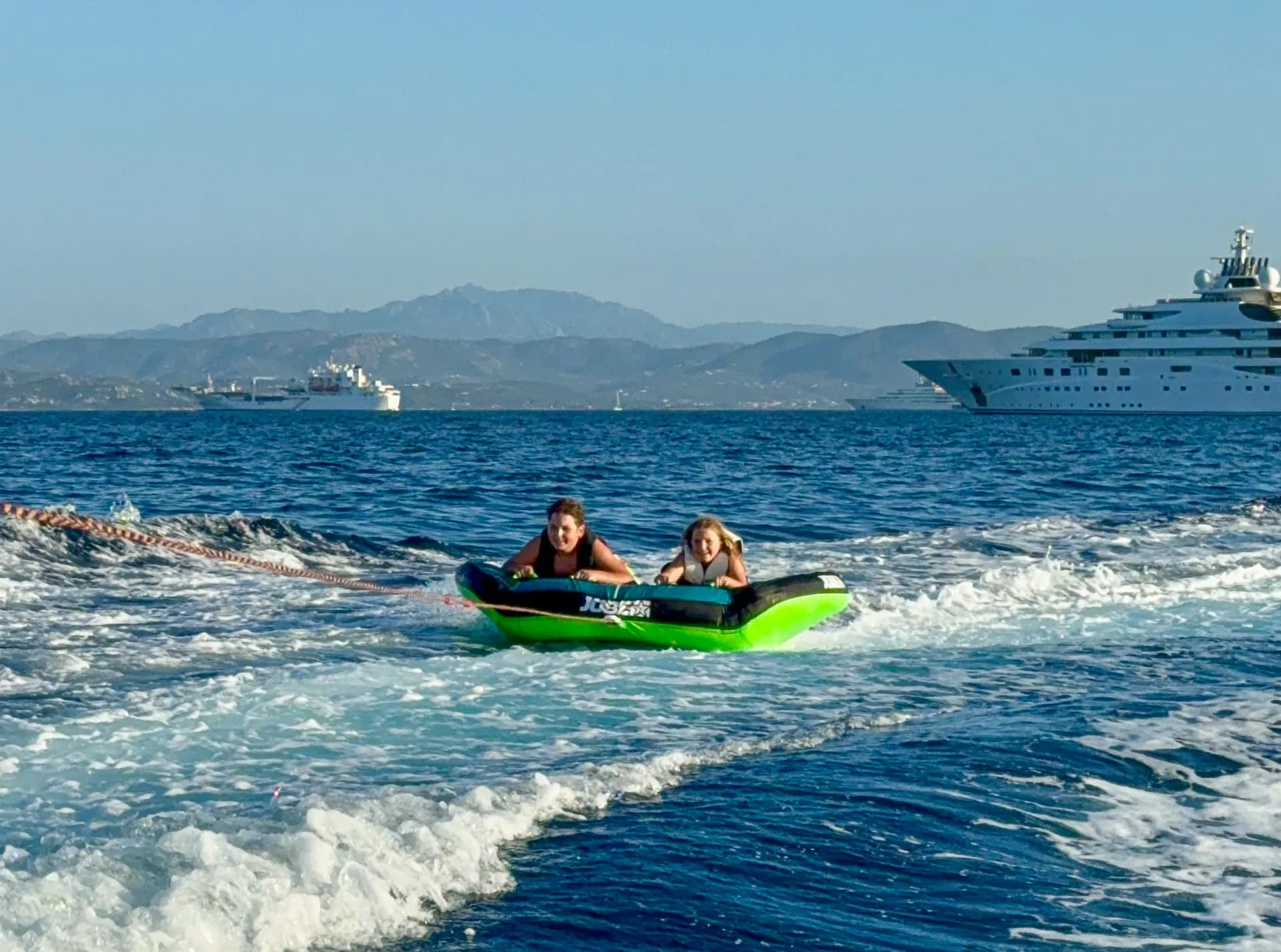 Zwei Kinder auf grünem Towreifen werden gezogen, eine Superyacht im Hintergrund